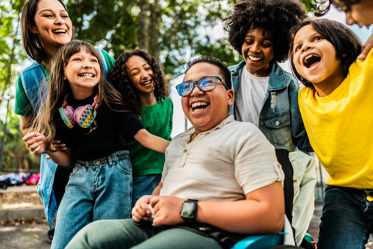Children and an adult in the outdoors smiling and having fun. One child is in a wheelchair, the others are standing around them.